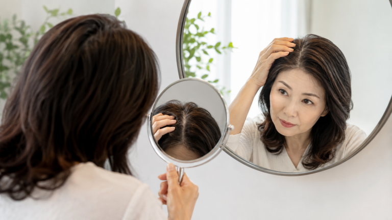거울을 보며 가르마와 정수리의 갱년기 탈모 초기 변화를 확인하는 중년 여성